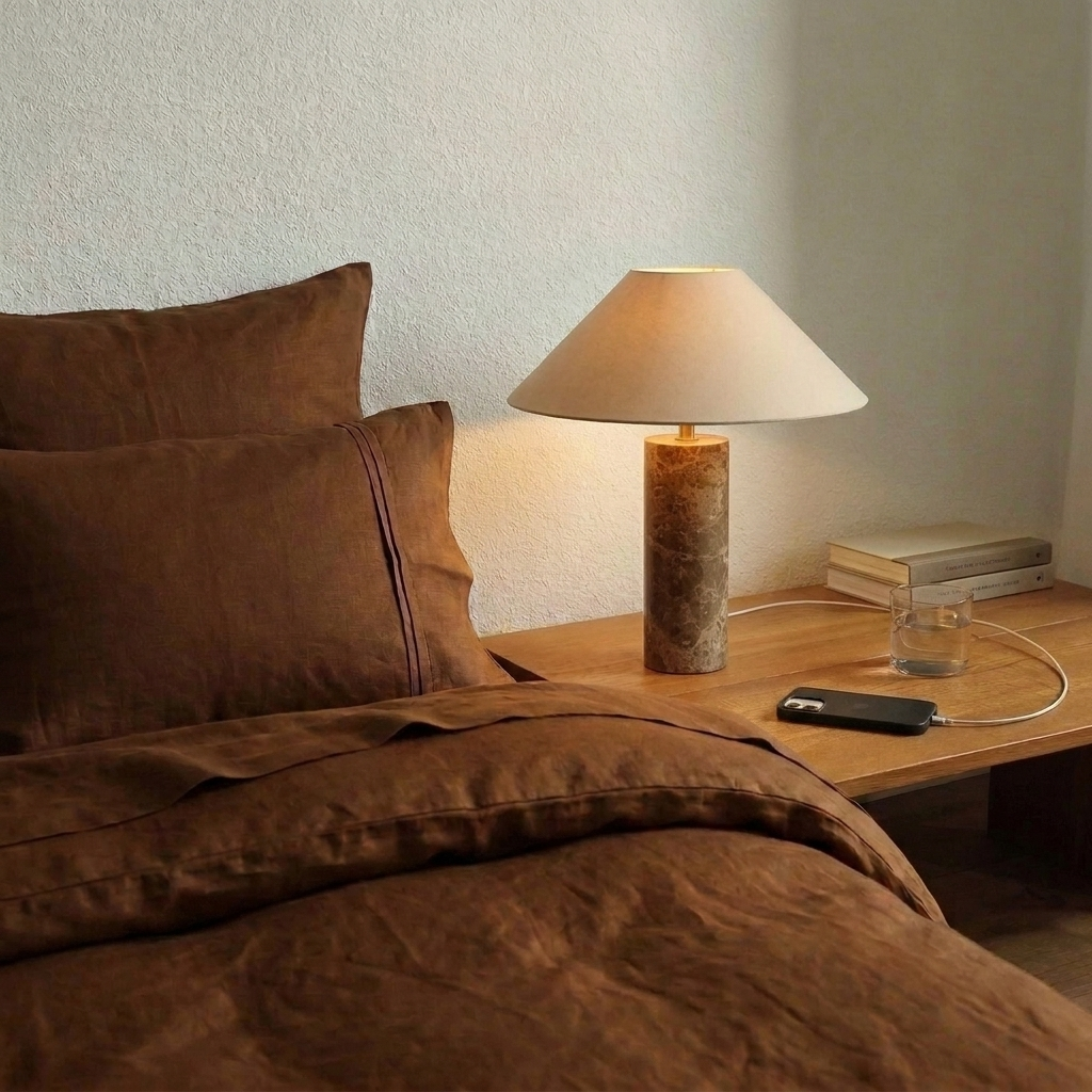 Lampe de chevet en marbre brun allumée sur une table de nuit en bois, à côté d'un lit en lin