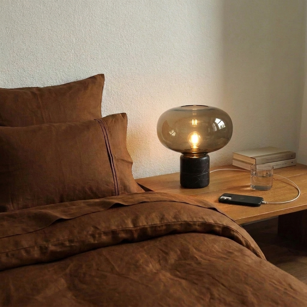 Lampe de chevet en marbre noir et verre cognac allumée sur une table de nuit en bois, à côté d'un lit en lin rouille
