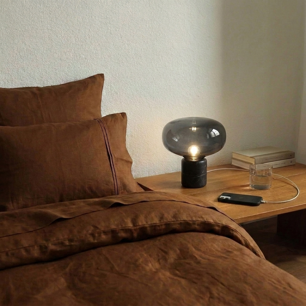 Lampe de chevet en marbre noir et verre fumé allumée sur une table de nuit en bois, à côté d'un lit en lin rouille