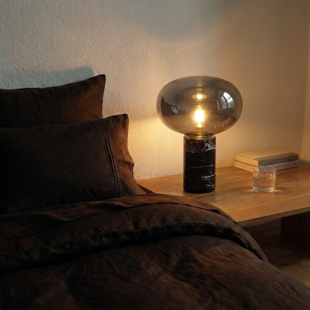 Lampe de chevet en marbre veiné et verre fumé allumée sur une table de nuit en bois, à côté d'un lit en lin chocolat