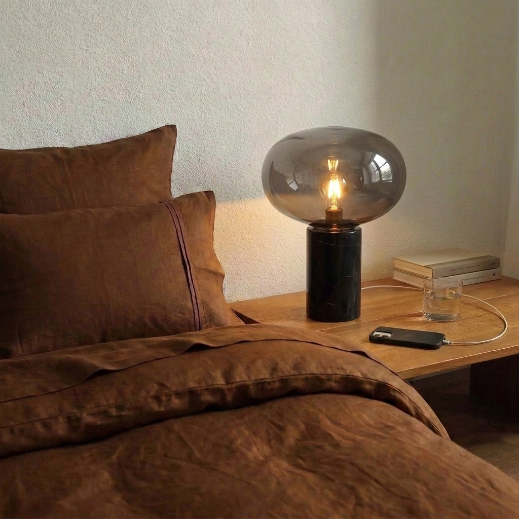 Lampe en marbre noir et verre fumé allumée sur une table de nuit en bois, à côté d'un lit en lin rouille
