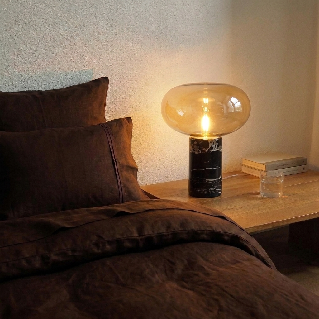 Lampe en marbre veiné et verre cognac allumée sur une table de nuit en bois, à côté d'un lit en lin chocolat